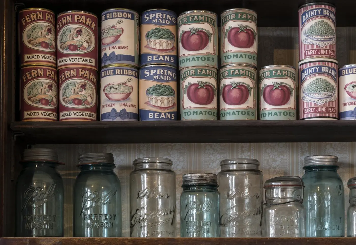 Two shelves of a dark wooden store cabinet, seen straight on in flat, even light. The upper shelf holds rows of canned food, their printed paper labels showing tomatoes, peas and beans. The lower shelf holds a row of glass canning jars, clear and pale blue-green, with metal lids and raised lettering in the glass. Striped wallpaper with a small floral pattern shows behind them.