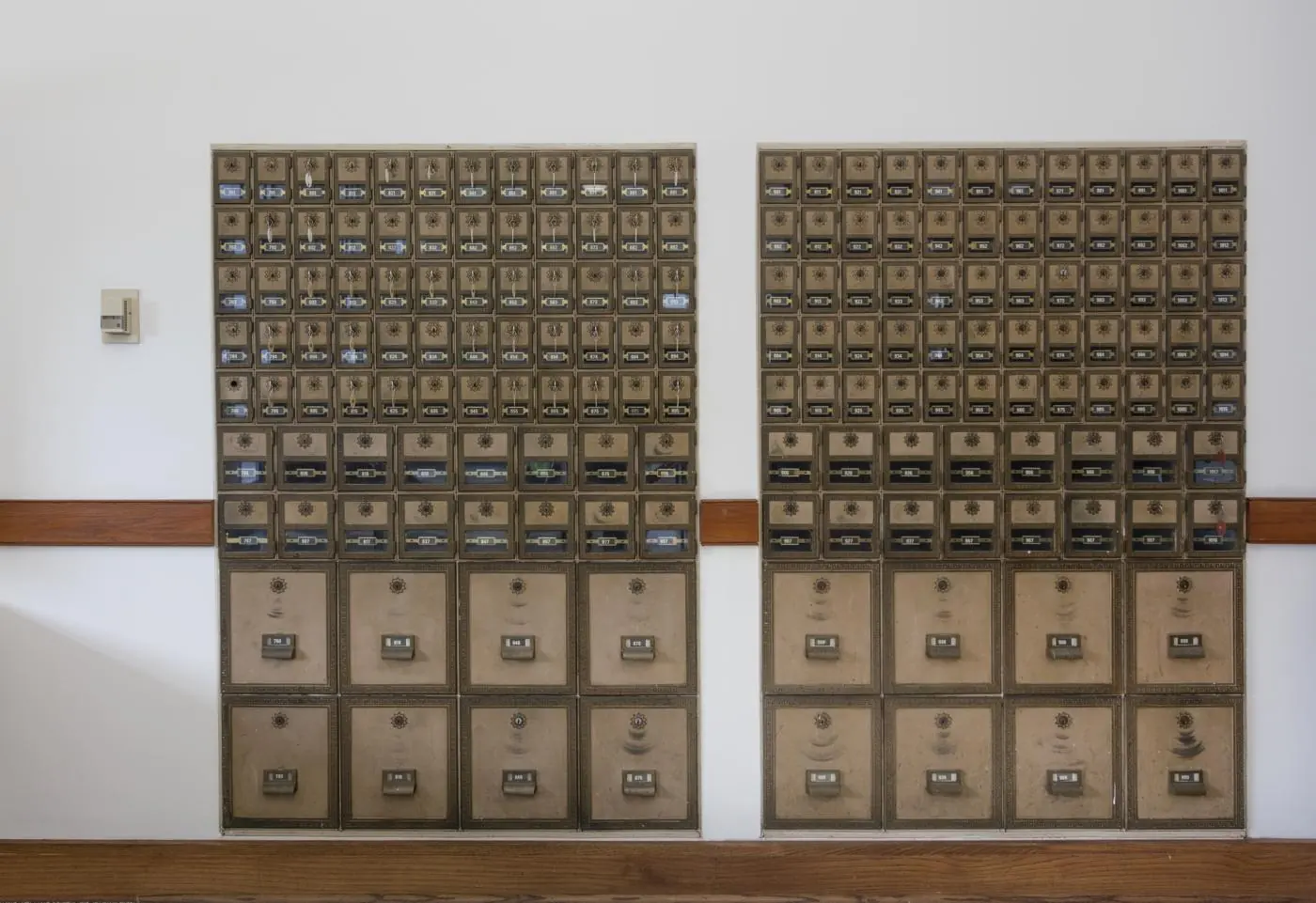 Two tall banks of antique brass post office boxes fill a pale wall, seen straight on in flat, even light. Rows of small doors with round dials and numbered plates run across the top two thirds; four large doors fill each of the bottom two rows. A dark wooden rail crosses the wall behind the banks, and a wooden board runs along the floor.
